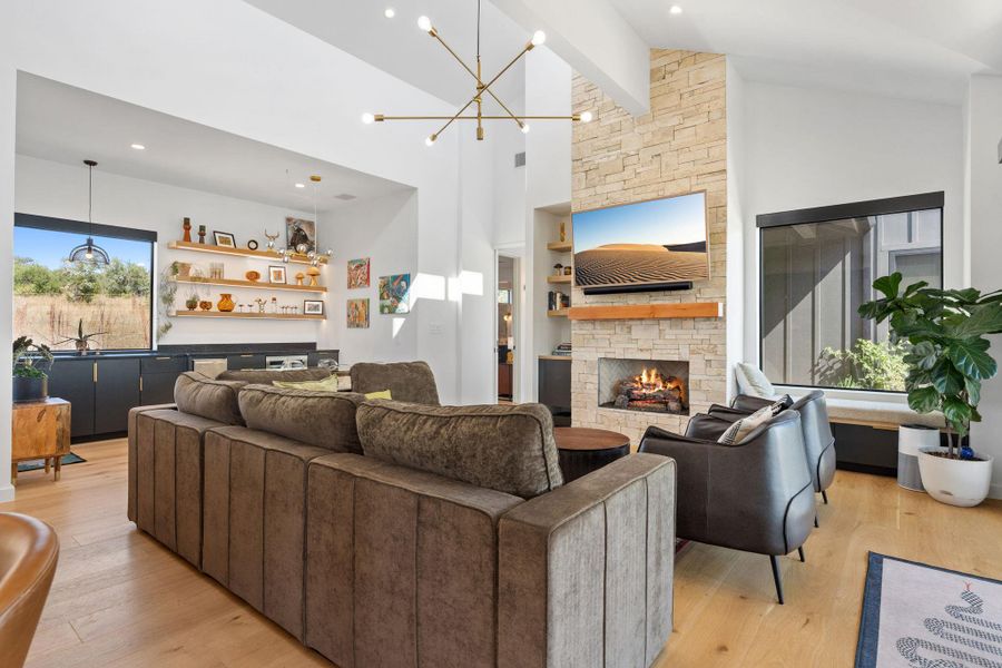 Spacious living area featuring a stacked stone fireplace with a wood mantle, high vaulted ceilings, large windows, wood-finish flooring, and a built-in wet bar with floating shelves