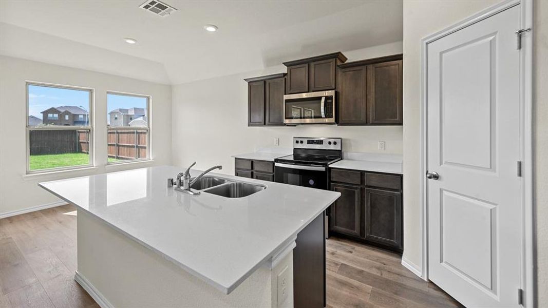 Kitchen featuring stainless steel appliances, dark wood finish cabinets, a center island with sink, light wood-type flooring, and light stone countertops
