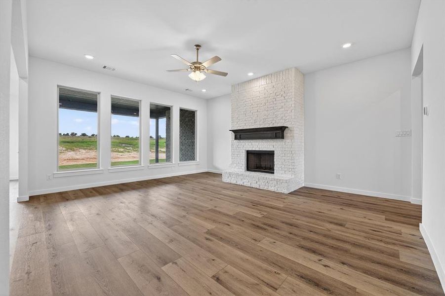 Spacious living area featuring expansive wood-finish flooring, a white-painted brick fireplace with a dark mantel, multiple large windows, recessed lighting, and a ceiling fan