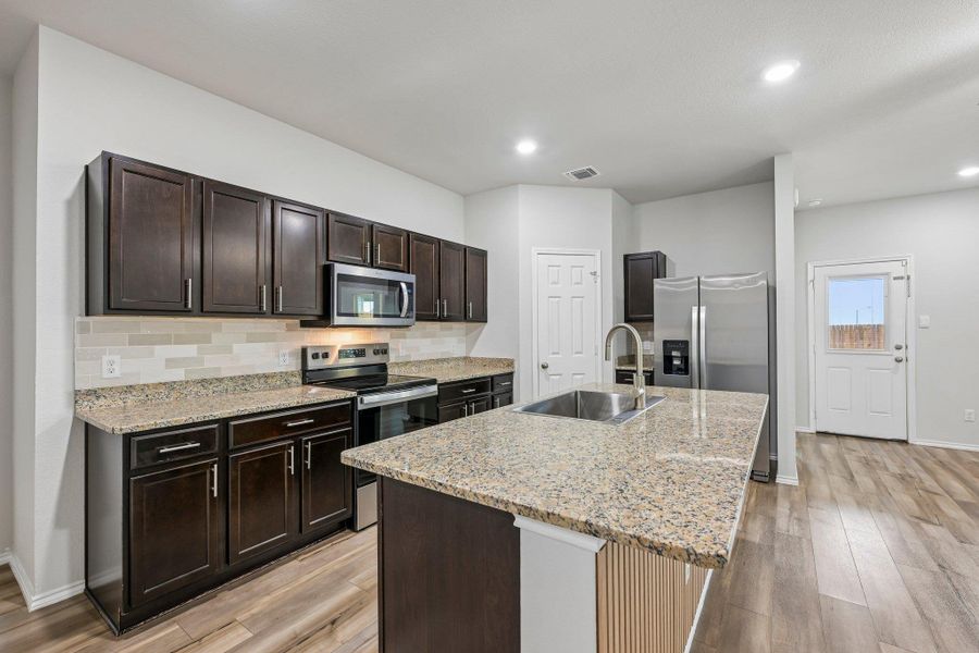 Kitchen featuring dark wood cabinetry, light granite countertops, stainless steel appliances, a tile backsplash, and wood-finish flooring
