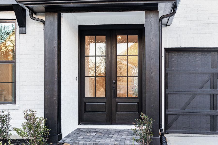 This striking modern farmhouse entryway features a bold black-framed, six-lite double front door and matching dark timber columns that create a sophisticated contrast against the crisp white brick exterior and grey cobblestone porch.