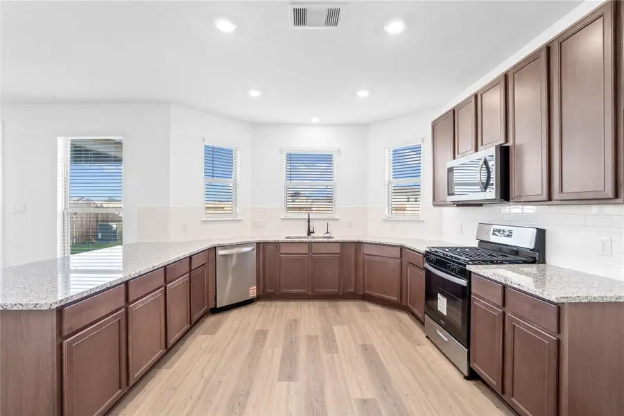 This spacious kitchen boasts soaring ceilings, Woodmont Dakota cabinets in a Smolder stain, Silestone Lagoon countertops, stainless steel Whirlpool appliances, a modern Daltile backsplash, recessed and pendant lighting, under-cabinet lighting, an extended breakfast bar countertop, and a pantry—all overlooking the expansive family room.