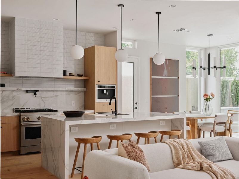 Open-concept kitchen and living area featuring a large center island with waterfall edge, integrated stainless steel range, light wood-finish cabinetry, and a full-height tile backsplash