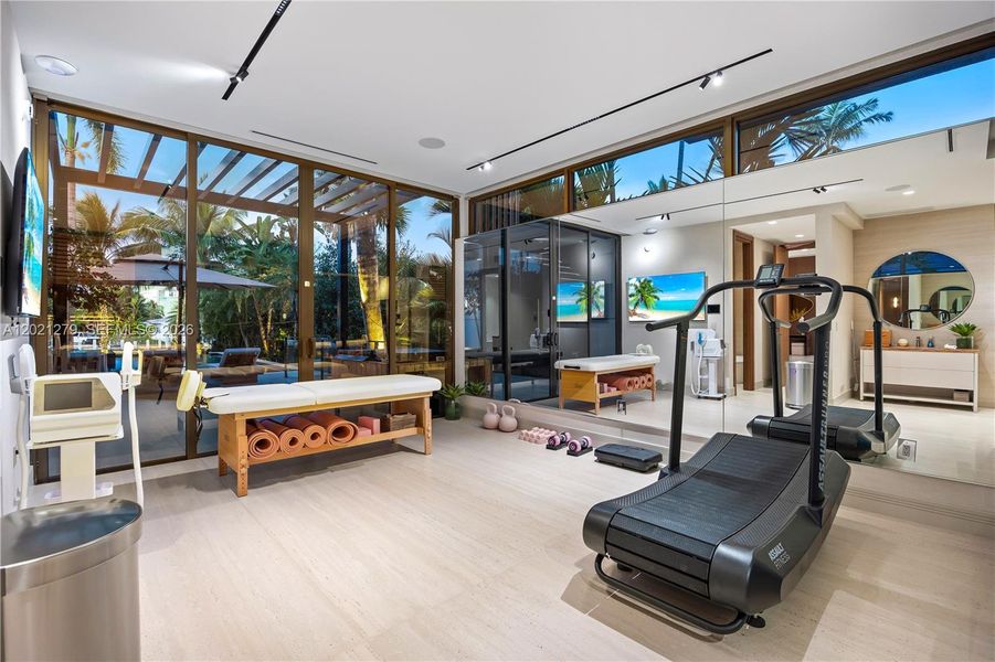 Full private wellness center with curved treadmill, massage table, yoga mats, kettlebells, wall-mounted TV, full-wall mirror, light wood floors, track lighting, clerestory windows, bronze-framed floor-to-ceiling glass walls, and direct pool and garden views.