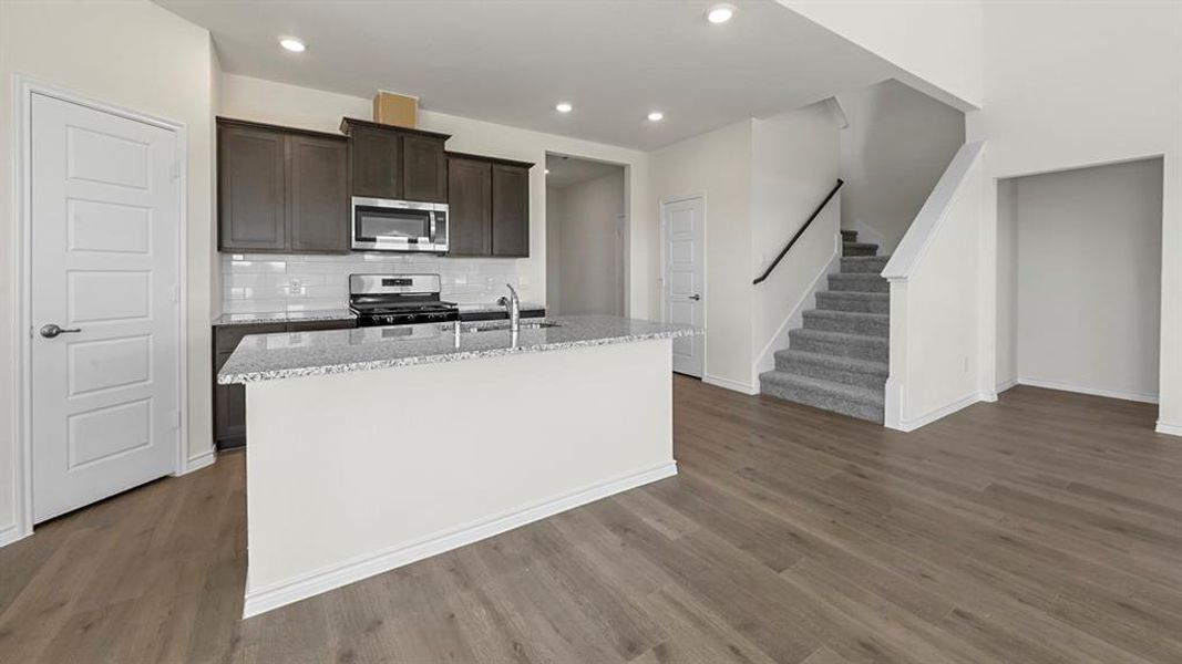 Kitchen with light stone counters, stainless steel appliances, an island with sink, dark brown cabinets, and dark wood-style flooring