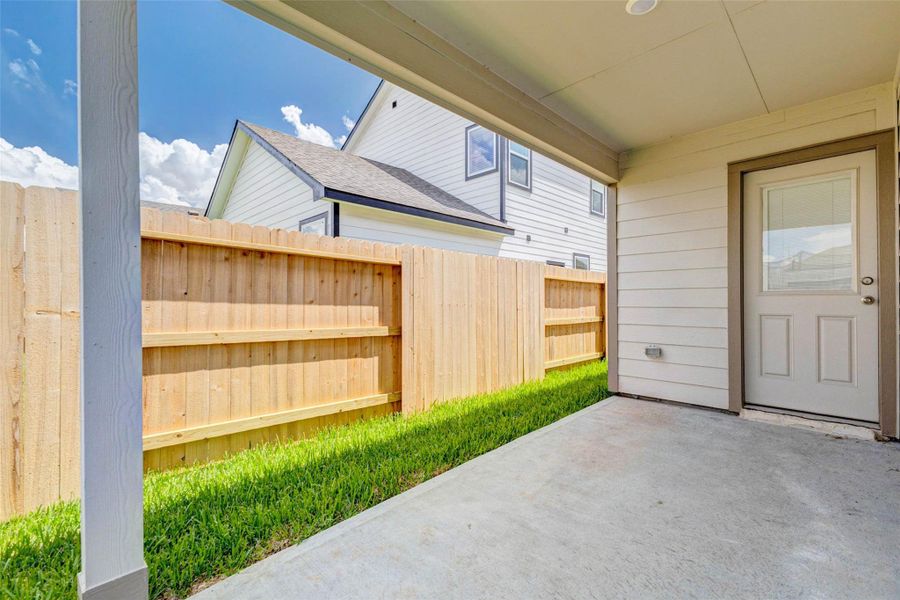 Covered back patio with concrete slab, rear entry door, privacy fence, and a narrow grassy side yard. This shaded outdoor space is ready for patio furniture, a grill, potted plants, or a cozy seating area, creating an easy transition from indoor living to private outdoor enjoyment.