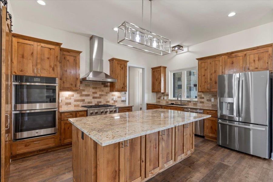 Kitchen featuring appliances with stainless steel finishes, wall chimney range hood, brown cabinets, a center island, and recessed lighting