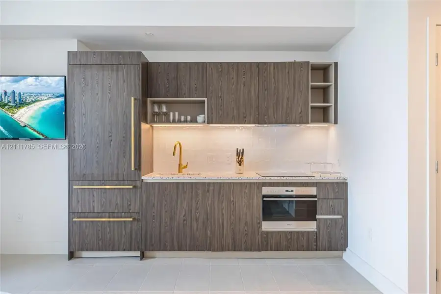 Kitchenette featuring dark wood-grain cabinetry, gold-toned hardware, stone-composite countertop, integrated refrigerator, and built-in wall oven