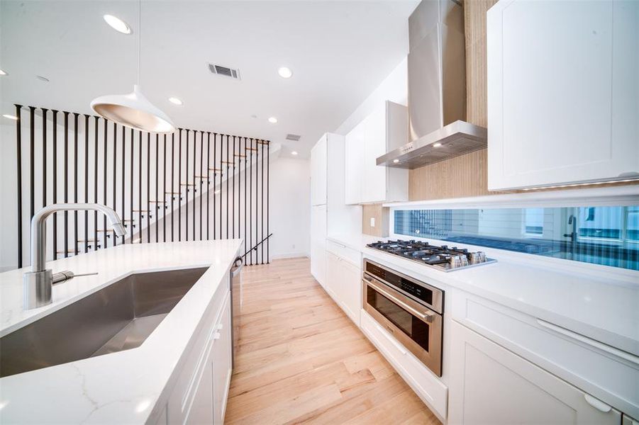 Kitchen with wall chimney range hood, light hardwood / wood-style flooring, stainless steel appliances, decorative light fixtures, and white cabinetry