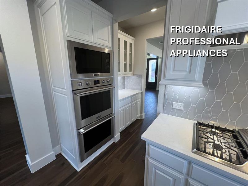 Kitchen featuring a stainless steel built-in double oven and microwave combination, white shaker cabinetry, a geometric tile backsplash, light-colored countertops, and dark wood-finish flooring Kitchen featuring a stainless steel built-in double oven and microwave combination, white shaker cabinetry, a geometric tile backsplash, light-colored countertops, and dark wood-finish flooring