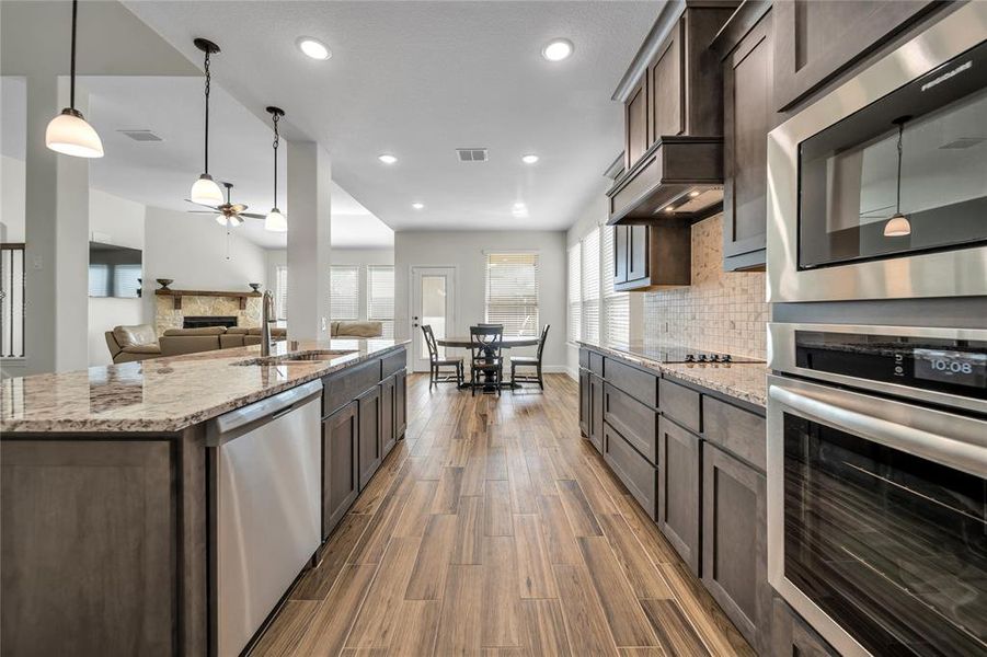 Kitchen featuring a fireplace, stainless steel appliances, pendant lighting, light stone counters, and dark brown cabinetry Kitchen featuring a fireplace, stainless steel appliances, pendant lighting, light stone counters, and dark brown cabinetry