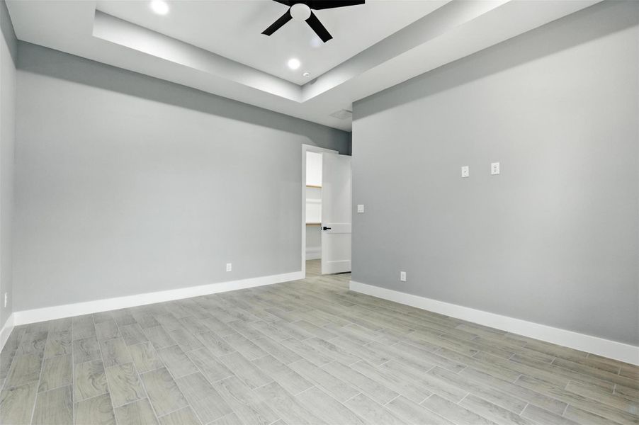 This room features modern gray walls, wood-look tile flooring, and a stylish tray ceiling with recessed lighting and a ceiling fan. A door opens to a walk-in closet, enhancing its functionality and appeal.
