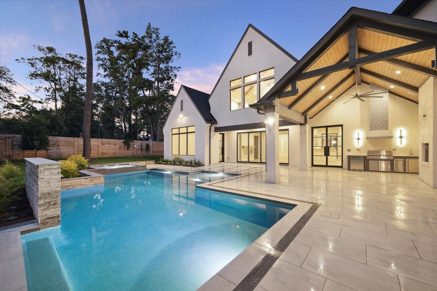 The backyard features a sleek swimming pool/spa, an outdoor kitchen, and a covered patio area. The house has large windows, contemporary architecture, and is surrounded by lush greenery, perfect for entertaining and relaxation.