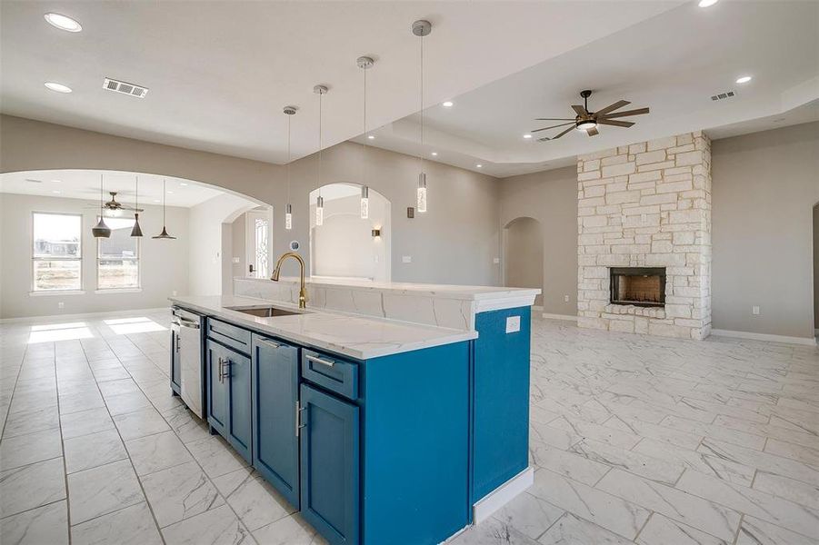 Kitchen with ceiling fan, sink, a center island with sink, a stone fireplace, and hanging light fixtures