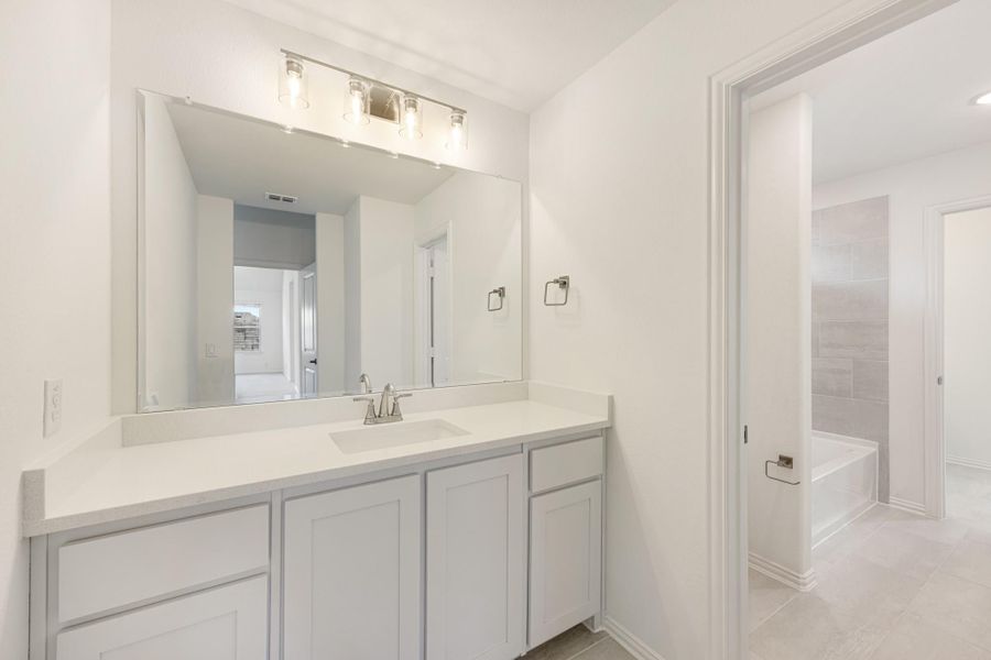 Bathroom with white vanity, quartz countertop, large mirror, and tiled soaking tub visible through doorway Bathroom with white vanity, quartz countertop, large mirror, and tiled soaking tub visible through doorway