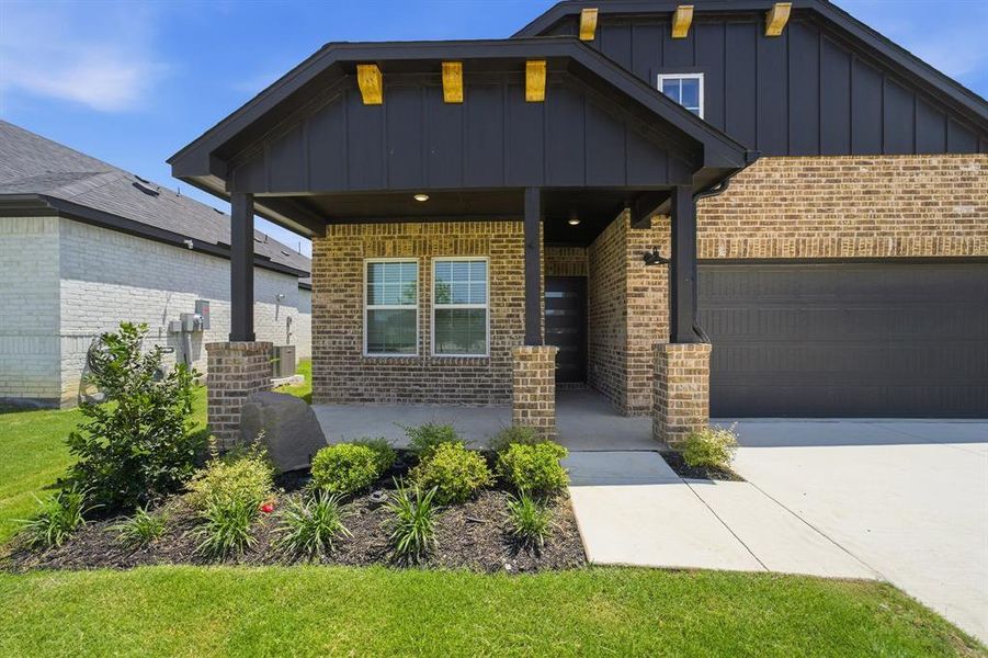 Brick facade home featuring a covered entryway, dark vertical siding, a dark garage door, a concrete driveway, and a landscaped garden bed