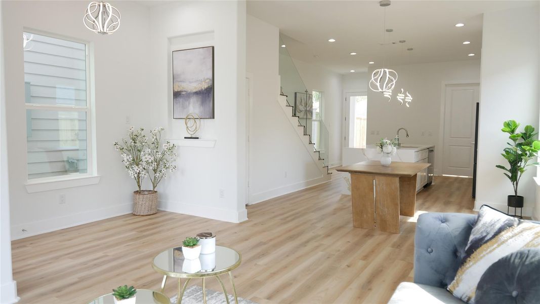 Bright and modern open-concept living area with light wood flooring and elegant lighting fixtures. Features a stylish kitchen island, contemporary staircase, and large windows for natural light. Ideal for comfortable living and entertaining. Bright and modern open-concept living area with light wood flooring and elegant lighting fixtures. Features a stylish kitchen island, contemporary staircase, and large windows for natural light. Ideal for comfortable living and entertaining.