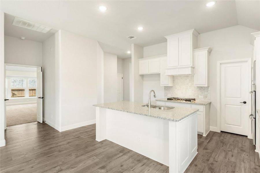Kitchen with white cabinets, granite island with sink, tile backsplash, and wood-look floors