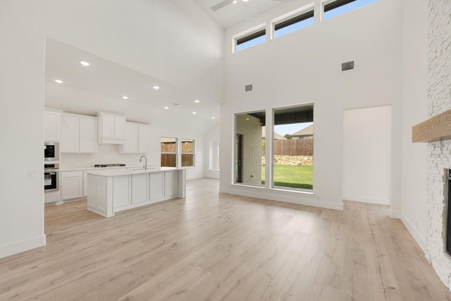 Open-concept living area with high ceilings, light wood floors, white kitchen with island, and stone fireplace