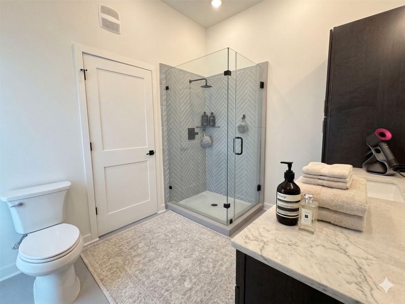 This modern bathroom features a glass-enclosed shower with herringbone tile, a sleek white toilet, a spacious vanity with a marble countertop, and contemporary fixtures, creating a clean and stylish space.