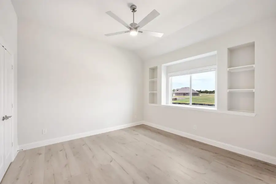 Empty bedroom with light wood floors, ceiling fan, vaulted ceiling, and built-in wall shelves beside a window