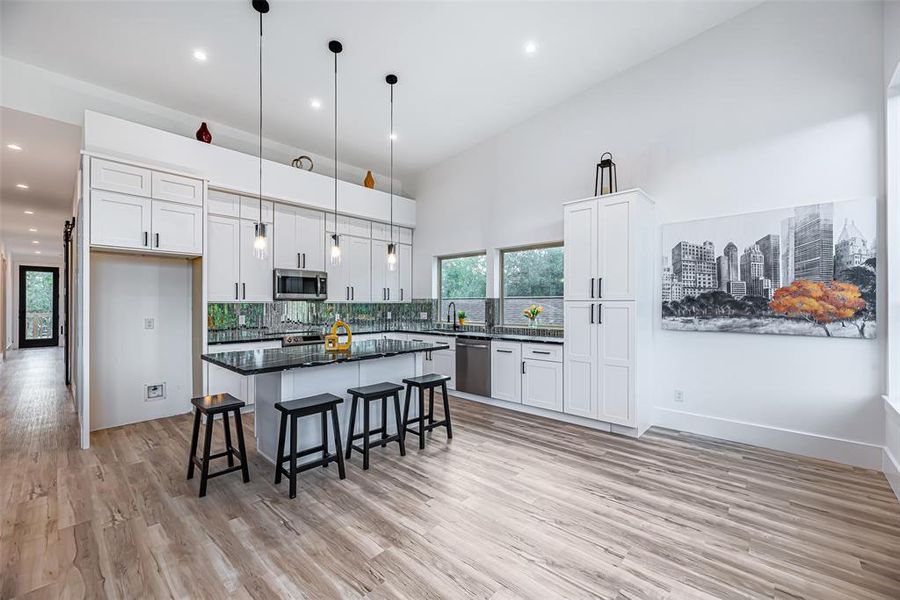 Bright and modern kitchen featuring white cabinetry, stainless steel appliances, a central island with bar seating, elegant pendant lighting, and sleek vinyl wood floors. Large windows provide natural light.