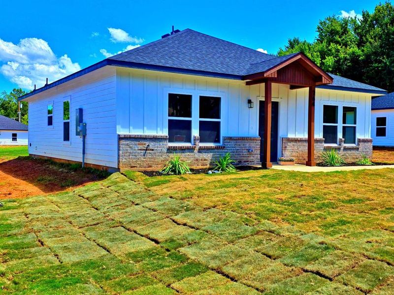 View of front of home featuring board and batten siding, a front lawn, a shingled roof, covered porch, and brick siding View of front of home featuring board and batten siding, a front lawn, a shingled roof, covered porch, and brick siding