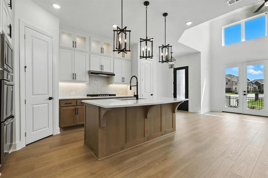 Kitchen featuring brown cabinetry, glass insert cabinets, decorative backsplash, hanging light fixtures, and dark wood-type flooring Kitchen featuring brown cabinetry, glass insert cabinets, decorative backsplash, hanging light fixtures, and dark wood-type flooring