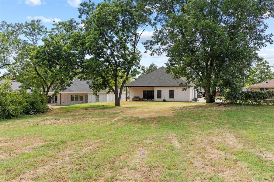 Expansive rear yard featuring mature trees and a white exterior home with a dark shingle roof and covered patio