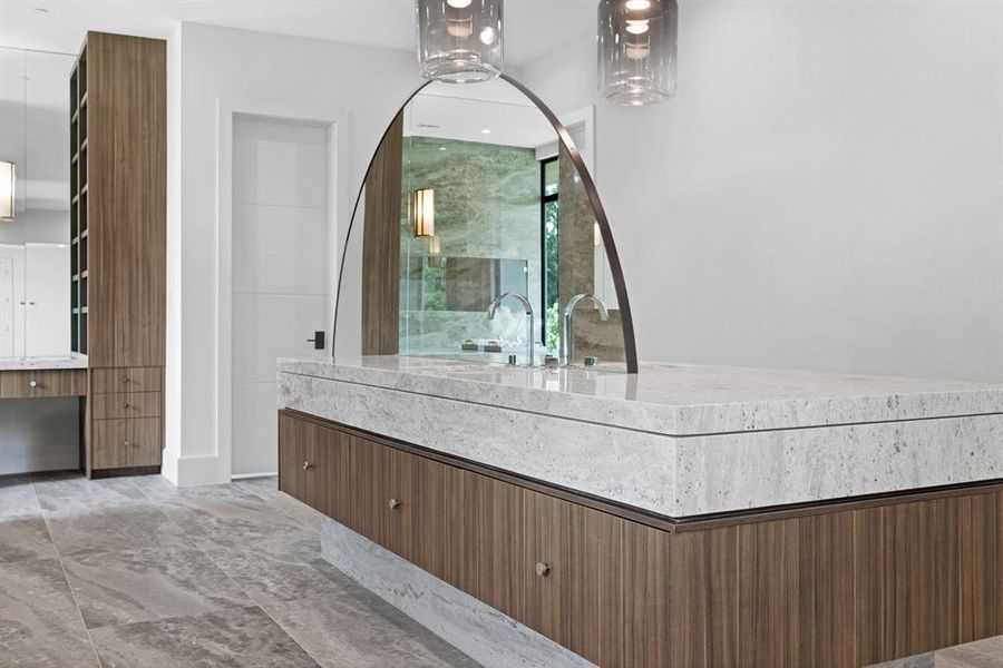 A custom dual-sided vanity offers ample storage and prep space for getting ready. A curvilinear mirror reflects natural light and creates a focal point in the space. Dual designer pendants illuminate the room and create an inviting atmosphere while keeping along with the spa-like color palette.