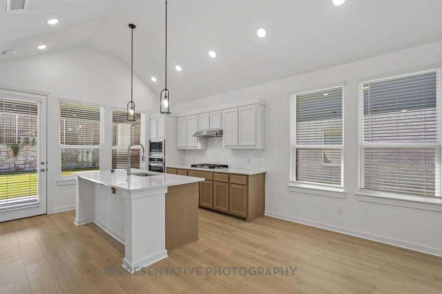 Kitchen featuring decorative backsplash, brown cabinetry, white cabinetry, decorative light fixtures, and an island with sink