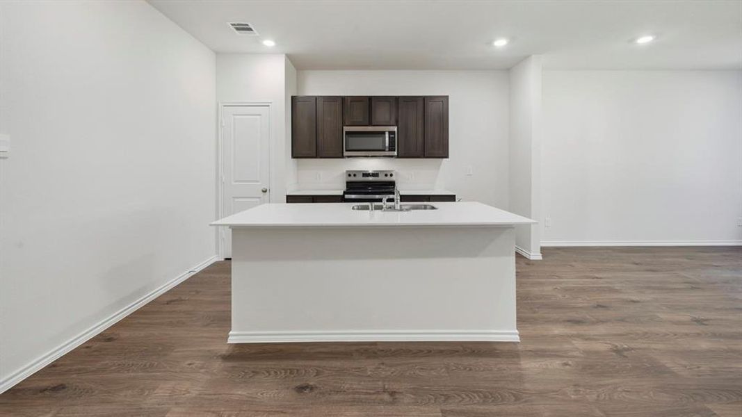 Kitchen featuring dark brown cabinetry, appliances with stainless steel finishes, a center island with sink, dark wood finished floors, and light stone counters