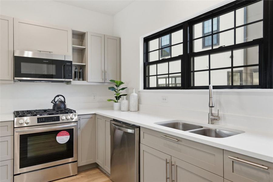 This kitchen features light gray cabinetry, a white countertop with double sink, sleek stainless steel appliances, and a large window that fills the space with natural light.