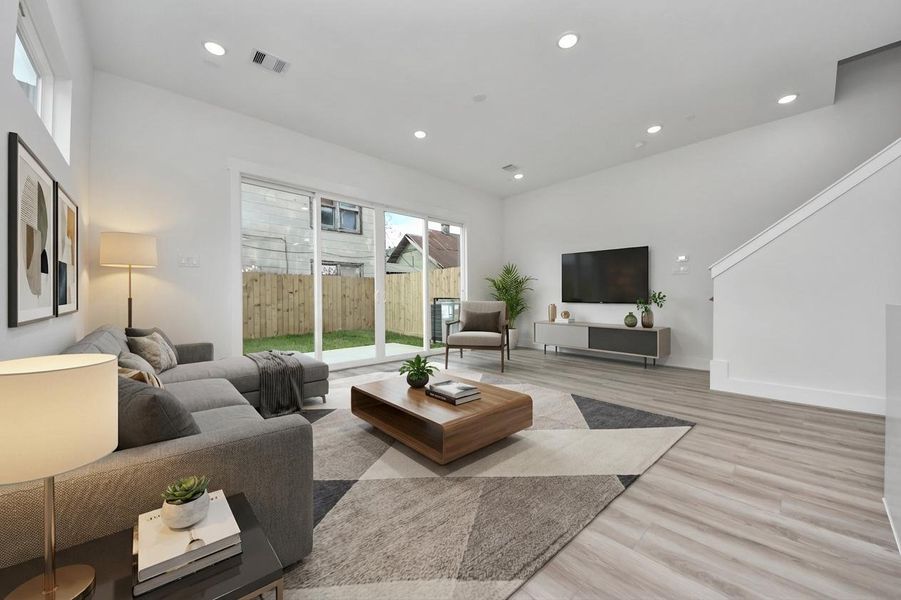 This photo shows a modern, bright living room with light wood flooring and neutral walls. It features a cozy sectional sofa, a stylish coffee table, and a wall-mounted TV. Large sliding glass doors open to a fenced backyard, providing natural light and outdoor access. The space feels open and inviting, ideal for relaxation and entertainment.
