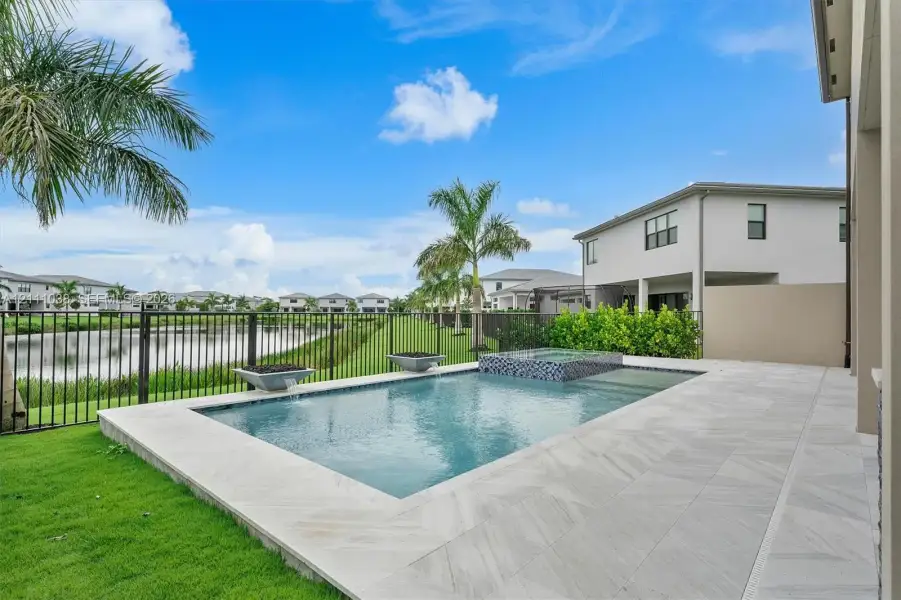 In-ground swimming pool with an integrated spa, stone-finish decking, manicured lawn, metal perimeter fencing, and water-side positioning