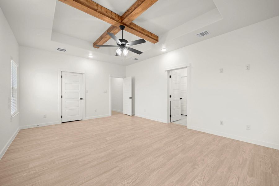 A spacious room features a tray ceiling with exposed wooden beams and a modern ceiling fan, all above a light-toned wooden floor.