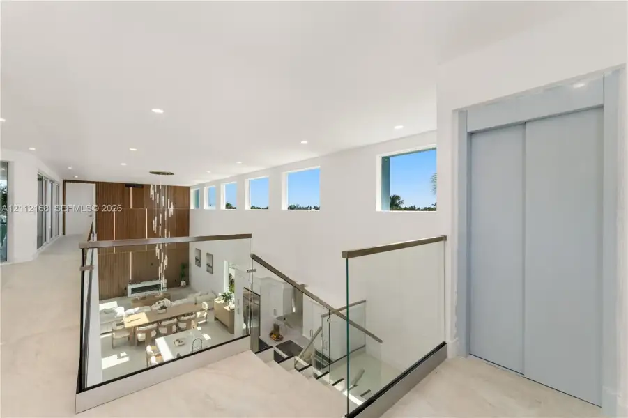 Open landing featuring large-format tile flooring, a glass-panel railing with a wood-finish handrail, a private elevator, recessed ceiling lighting, and a series of high-placement fixed windows Open landing featuring large-format tile flooring, a glass-panel railing with a wood-finish handrail, a private elevator, recessed ceiling lighting, and a series of high-placement fixed windows
