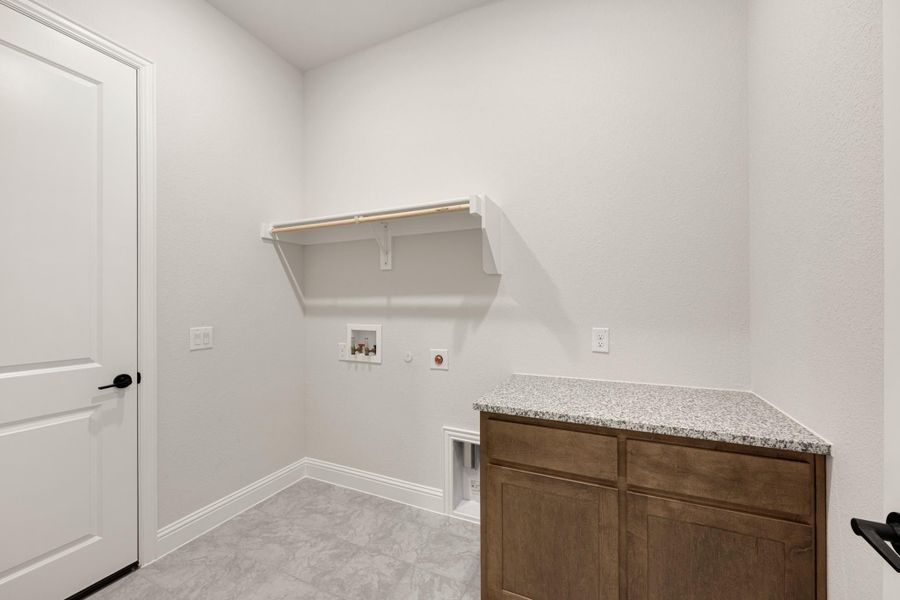 Laundry room with wood cabinet, granite countertop, wall shelf, and washer and dryer hookups on tile floor. Laundry room with wood cabinet, granite countertop, wall shelf, and washer and dryer hookups on tile floor.