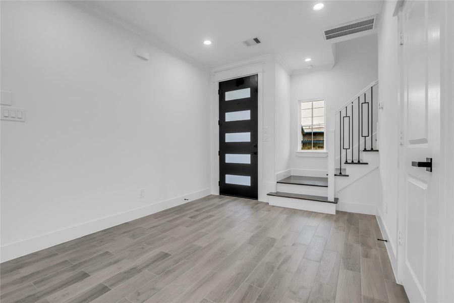 This modern entryway features sleek wood flooring, a striking black front door with frosted glass panels, and a staircase with contemporary railings. The space is bright and welcoming, offering a clean, minimalist aesthetic.