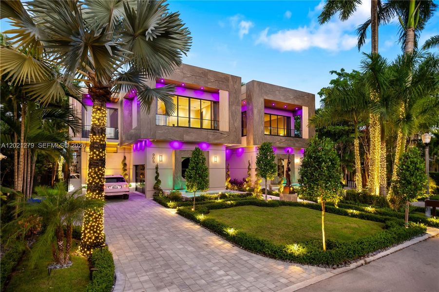 Modern two-story luxury home with stone façade, paver driveway, covered porte-cochère, manicured tropical landscaping, royal palms, and programmable LED lighting.