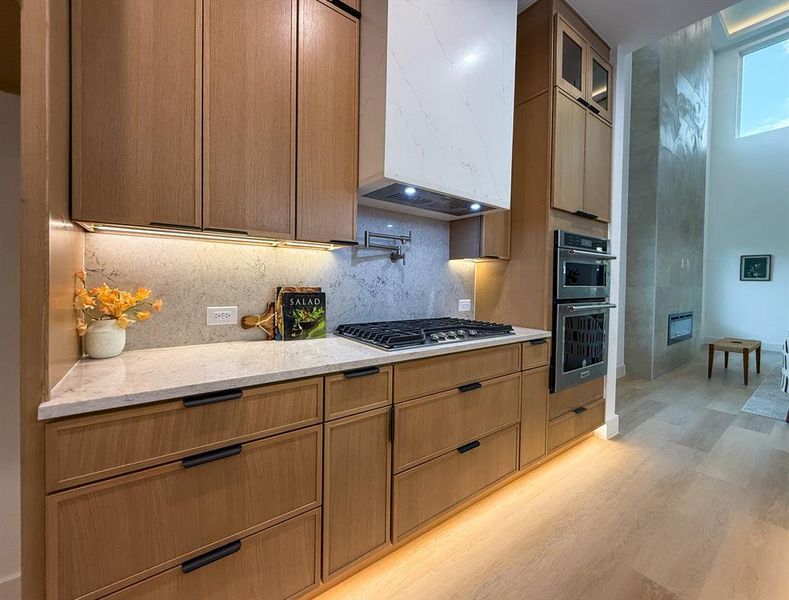 Kitchen with light stone countertops, backsplash, glass insert cabinets, light wood-style flooring, and extractor fan