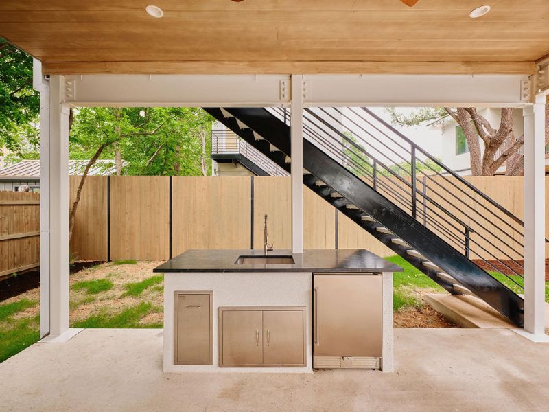 Covered patio featuring an outdoor kitchen island with a dark countertop, integrated sink, and stainless steel appliances