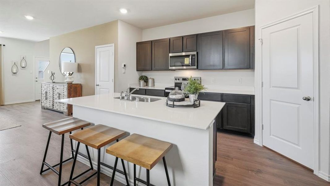 Kitchen with dark wood-style flooring, appliances with stainless steel finishes, a kitchen island with sink, a breakfast bar area, and light stone countertops