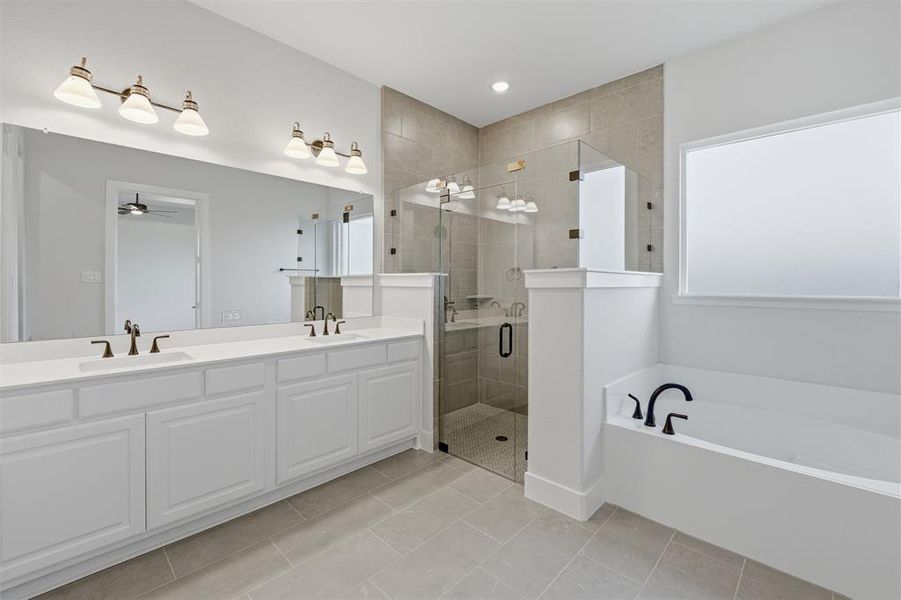 Bathroom featuring a double vanity with white cabinetry, a glass-enclosed shower with tile surround, a freestanding bathtub with dark fixtures, and light-colored tile flooring