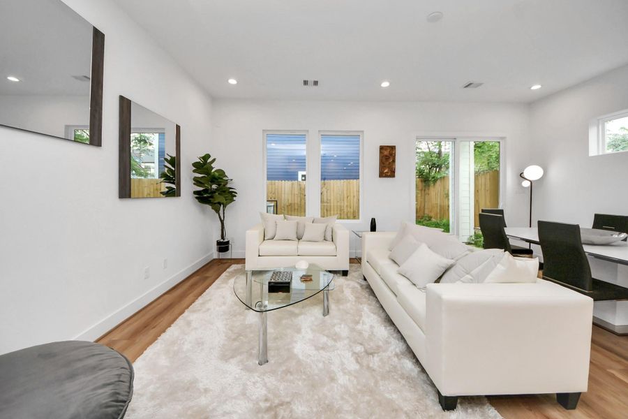This inviting living room boasts modern elegance with its bright, open layout, chic white sofas, and warm wooden floors, seamlessly connecting to a serene outdoor space.