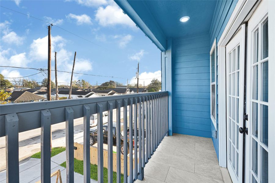 This photo showcases a small balcony with blue siding and white French doors, offering a view of a residential street. The area is well-lit and features a railing for safety, providing a cozy outdoor space.