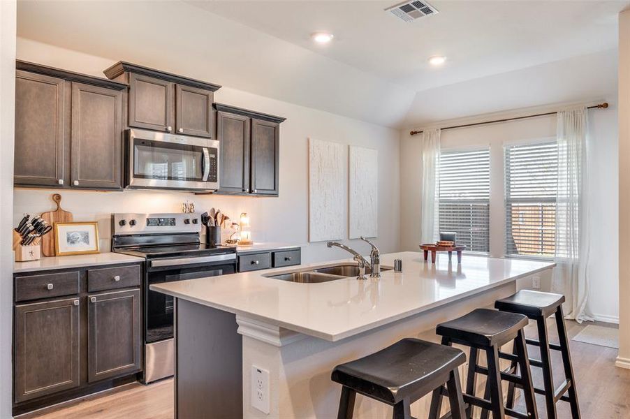 Kitchen featuring stainless steel appliances, dark brown cabinetry, light wood finished floors, a center island with sink, and light stone counters