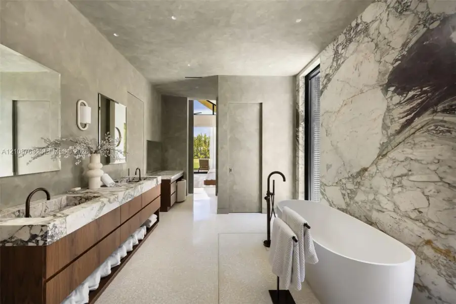 Custom vanity with integrated stone sinks and wood-finish cabinetry, freestanding soaking tub, expansive stone-slab feature wall, and recessed ceiling lighting