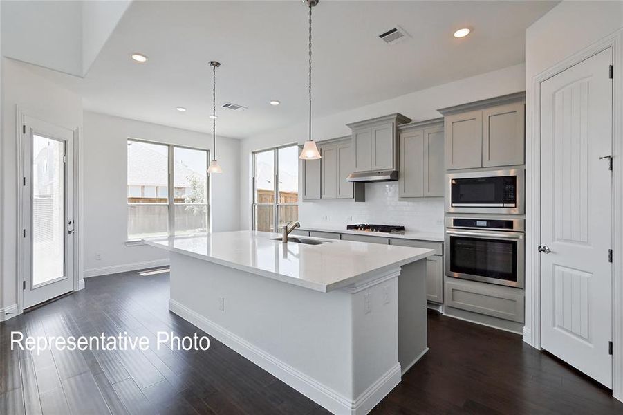 Spacious kitchen featuring a large central island with sink, light gray cabinetry, stainless steel appliances, and wood-finish flooring
