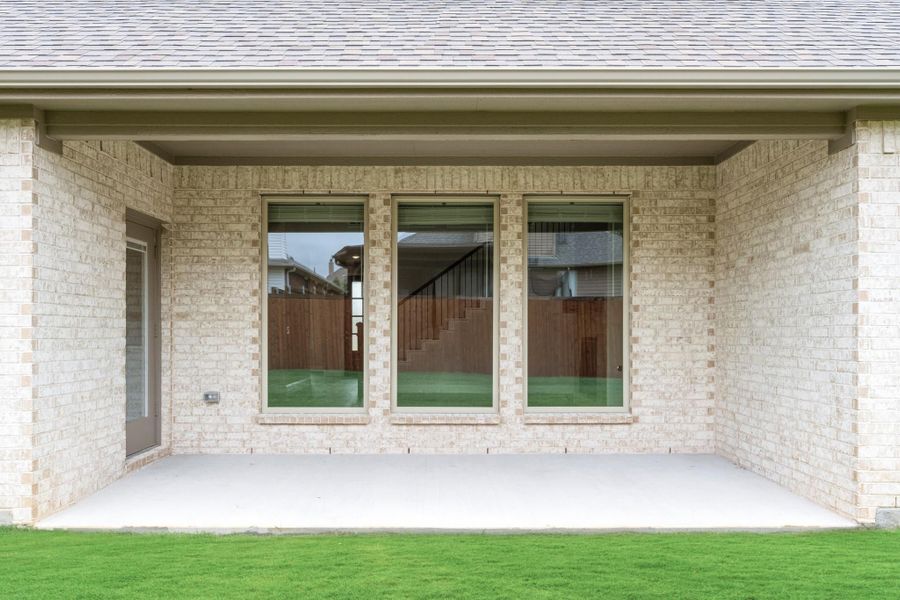 Covered back patio with concrete slab, light brick exterior, three large windows, and green lawn